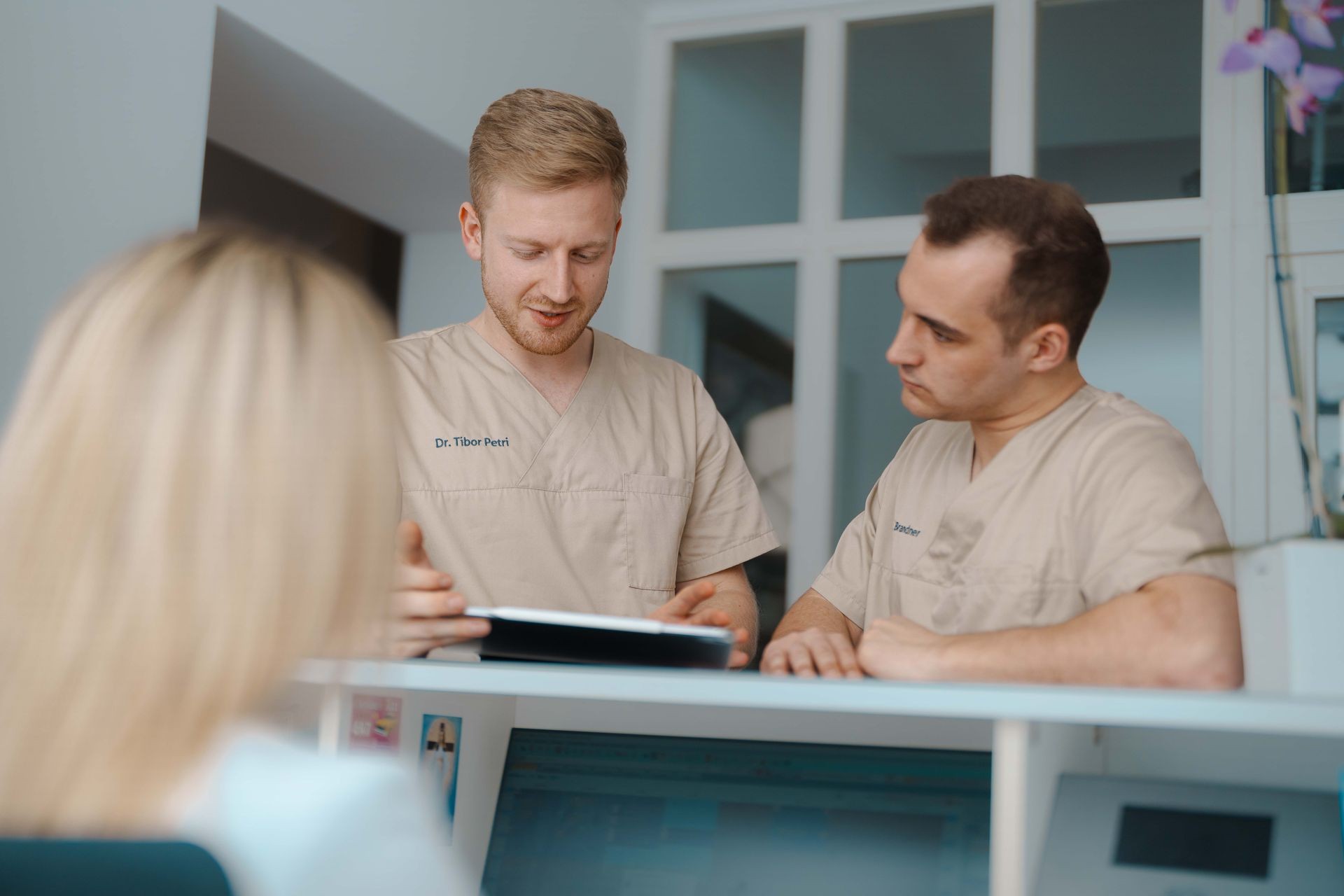 Dr. Tibor Petri und Dr. Karl Brandner in beigen Kitteln besprechen Unterlagen am Schalter, Monitor im Vordergrund